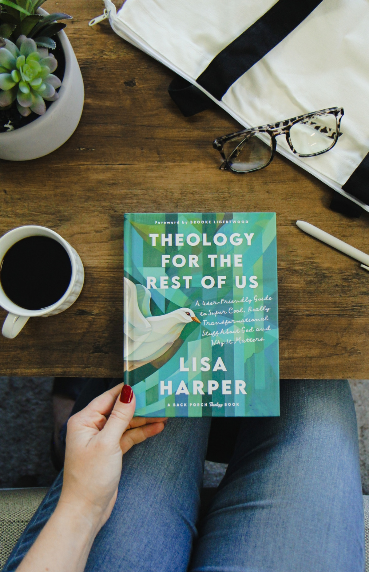 A copy of "Theology for the Rest of Us" being help by someone on the side of a brown table, next to a full coffee mug and a pair of glasses.