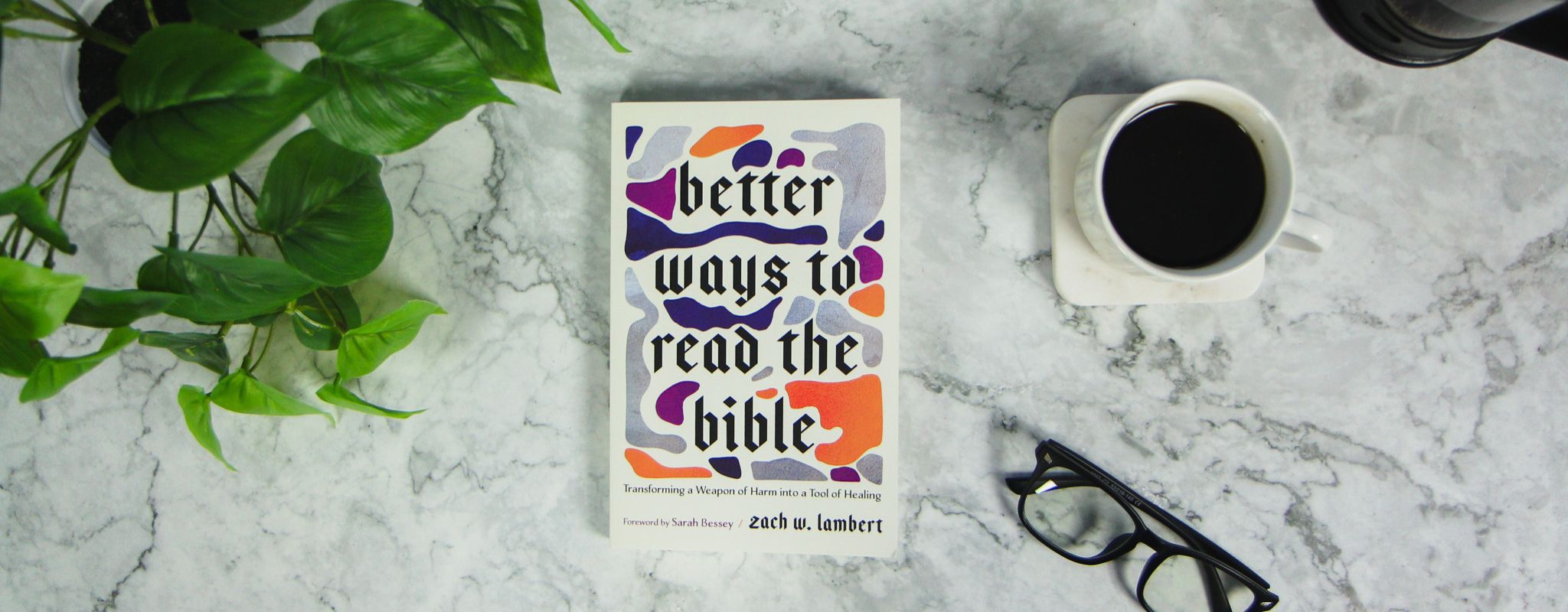 Image of the book "Better Ways to Read the Bible: Transforming a Weapon of Harm into a Tool of Healing" by Zach W. Lambert, on a white marble countertop next to a full coffee cup, a pair of glasses, and a green houseplant.
