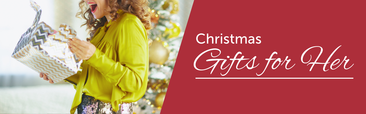 Christmas Gifts for Her. In the image, a woman excitedly opens a wrapped present, with a Christmas tree with gold ornaments in the background.