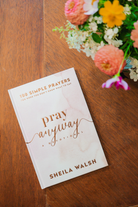 The cover of "Pray Anyway" by Sheila Walsh on a wooden table next to a bouquet of red and yellow flowers.