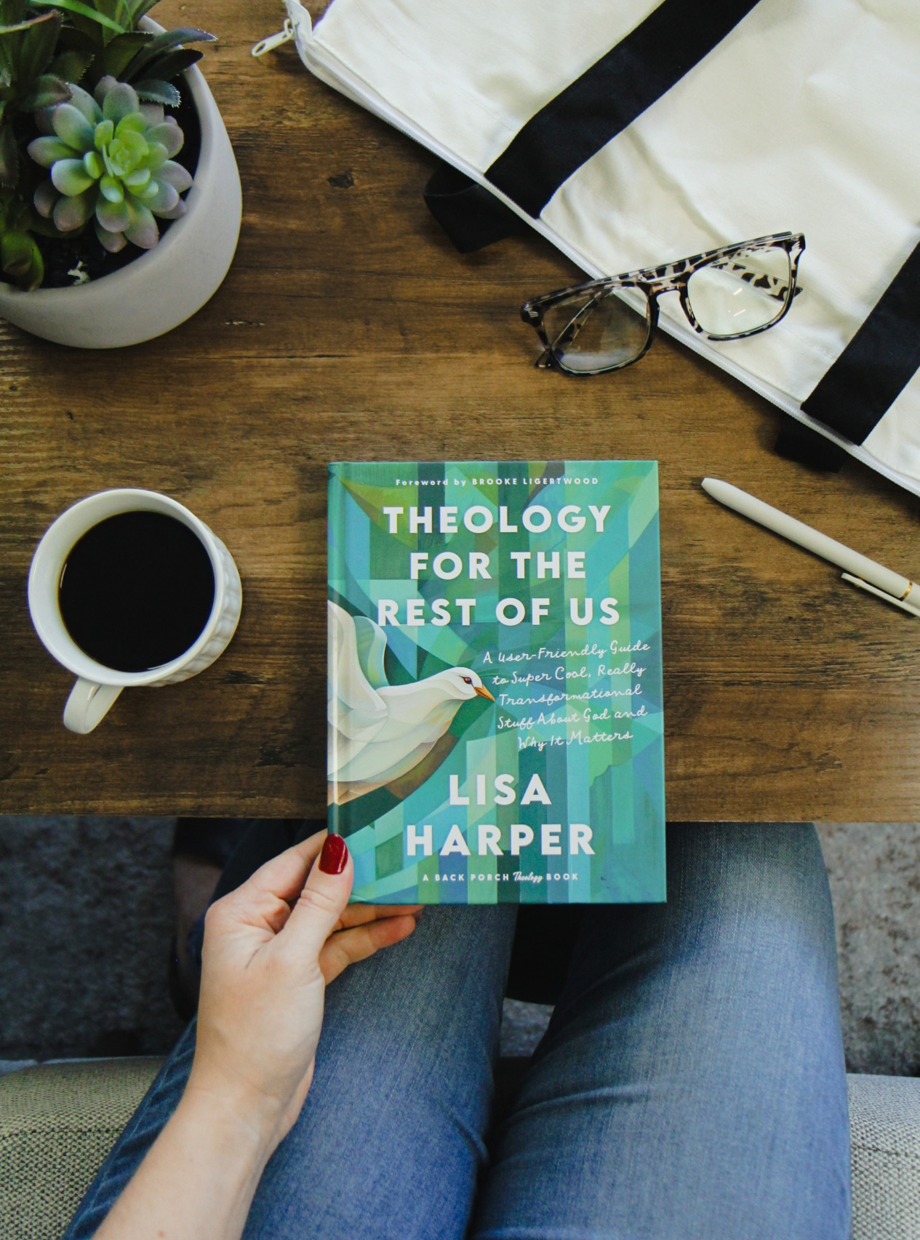 A copy of "Theology for the Rest of Us" being help by someone on the side of a brown table, next to a full coffee mug and a pair of glasses.