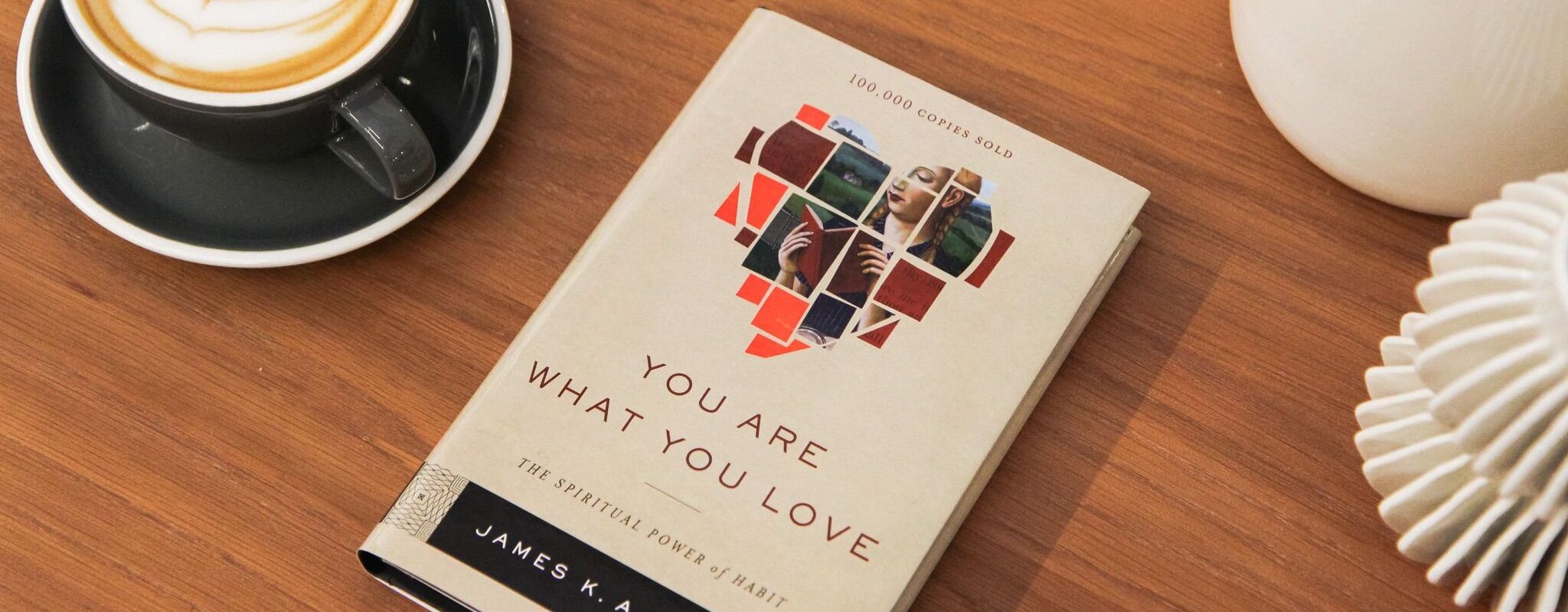 Image of the book "You Are What You Love: The Spiritual Power of Habit" by James K.A. Smith on a brown wooden table next to a full cup of coffee on a saucer.
