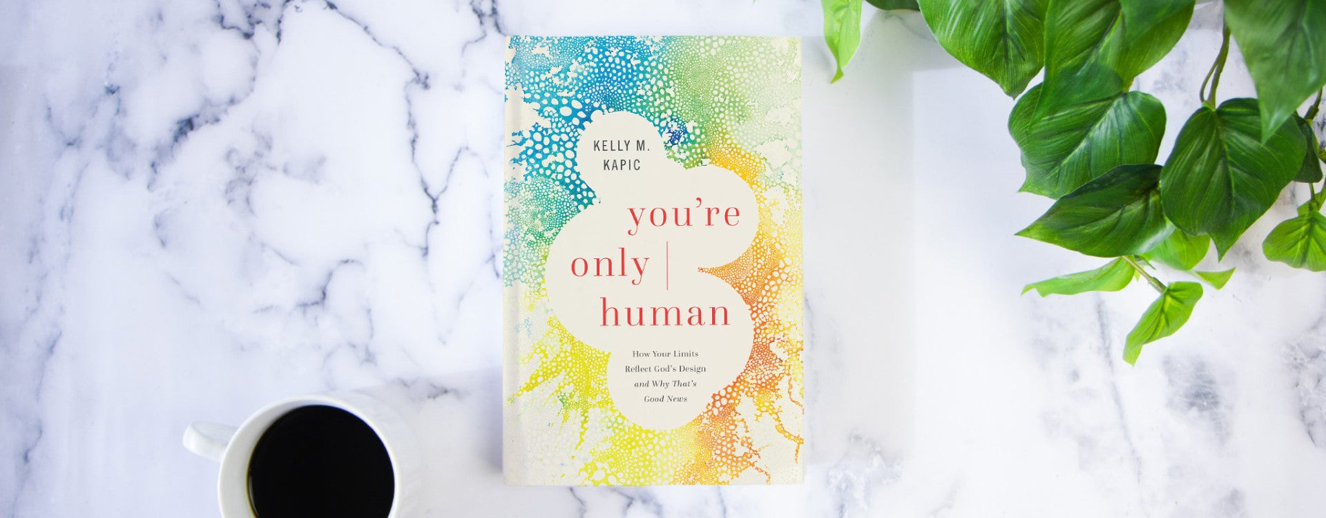Image of the book "You're Only Human: How Your Limits Reflect God's Design and Why That's Good News" by Kelly M. Kapic, on a white marble countertop next to a full cup of coffee and a green houseplant.